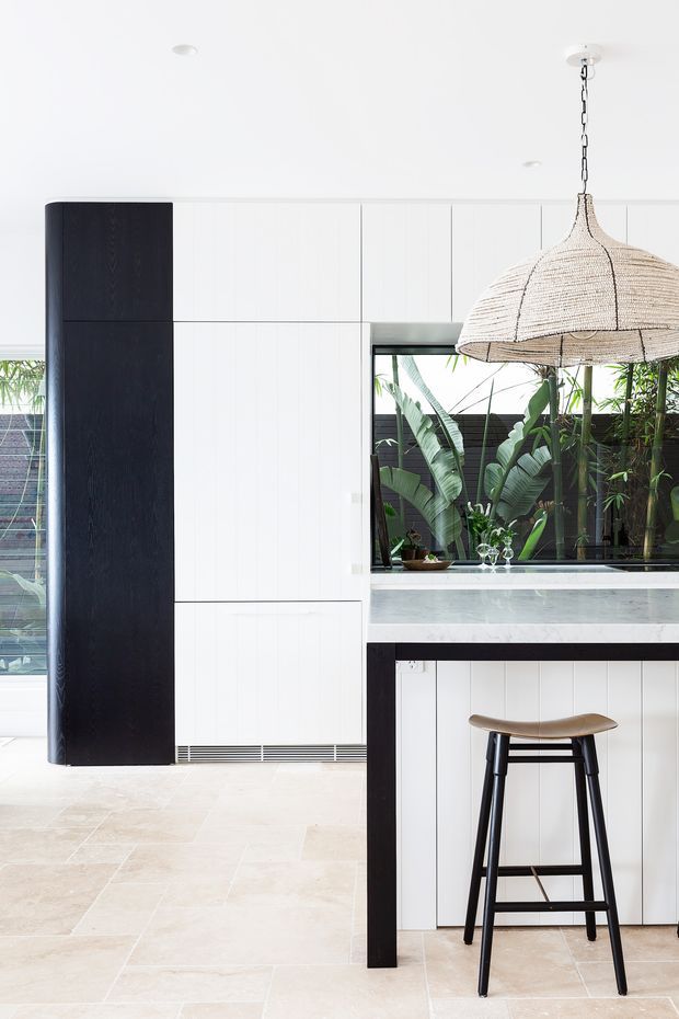 Bright contemporary kitchen featuring full-height white vertical-panel cabinetry, a contrasting dark timber pantry or cabinet column, pale stone benchtops, and a dark-framed island or peninsula. A woven rattan pendant light hangs above the island, with a timber-and-black bar stool in the foreground. Large windows reveal lush tropical planting, and the floor is finished in large pale stone tiles.