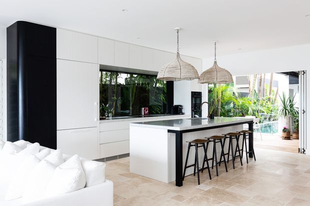 Bright contemporary open-plan kitchen with extensive white flat-panel cabinetry, a large white island with dark stone benchtop, black accents, and four bar stools. Woven pendant lights hang above the island, while full-height opening and glazing connect the kitchen to a lush outdoor area. A white sofa is visible in the foreground.