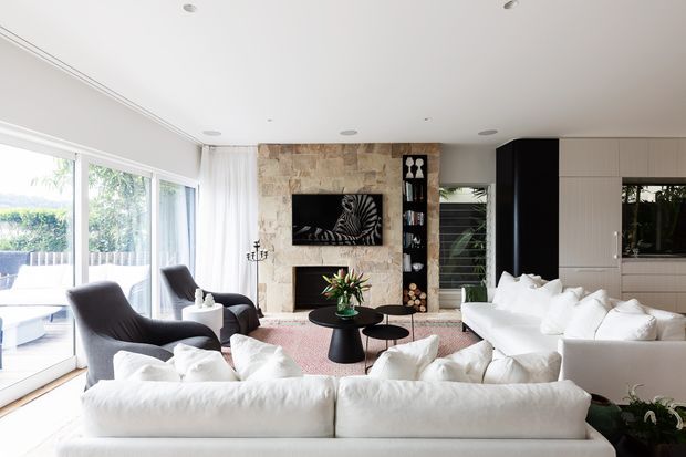 Bright, spacious contemporary living room with large white sectional sofas and dark lounge chairs arranged around low coffee tables. The room features expansive floor-to-ceiling glazing, a stone-clad fireplace wall with television, dark built-in shelving, and integrated light-toned cabinetry. A glimpse of an outdoor area is visible through the glazing.