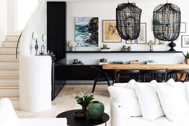 Open-plan contemporary living and dining area with a bright white sectional sofa in the foreground, a long timber dining table with black chairs, and a black built-in cabinetry and shelving wall displaying artwork and decor. A sculptural curved white kitchen island and staircase are visible to the left, with oversized woven pendant lights above the dining area and pale stone-look flooring throughout.