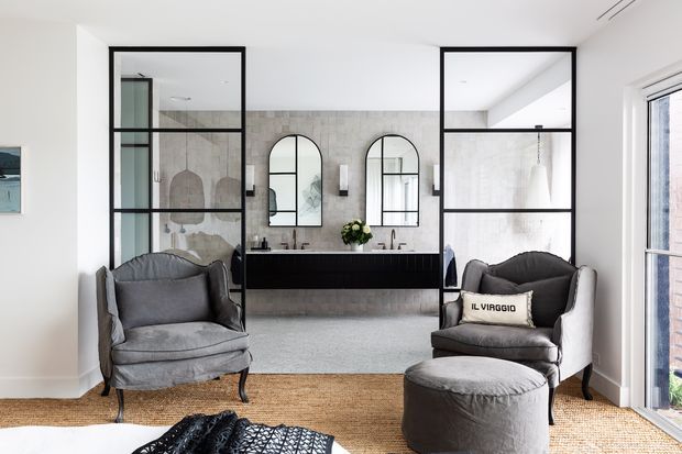 Bright, refined bathroom viewed from an adjoining sitting area, with black-framed internal glass partitions, a long dark floating vanity, twin basins, arched mirrors, pale stone or concrete-look wall tiling, and grey upholstered lounge chairs in the foreground. Natural light enters through full-height glazing at the right.
