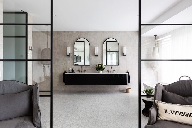 A refined open-plan bathroom viewed through black-framed glass partitions, with a floating dark vanity, twin vessel basins, arched mirrors, pale stone-look tiled walls, and terrazzo-style flooring. Upholstered lounge chairs are visible in the foreground, creating a connection to an adjoining living area. The space is bright, contemporary, and monochromatic with strong black accents.