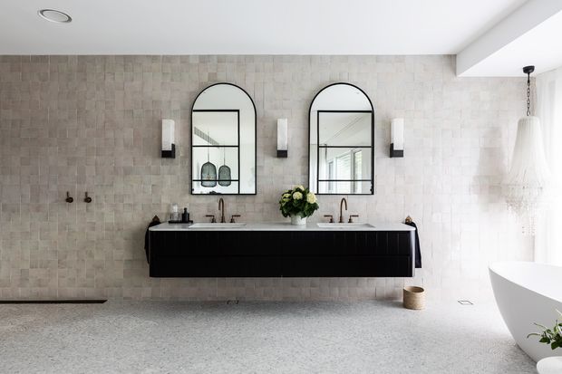 A bright, contemporary bathroom featuring a long floating black double vanity with twin basins, arched black-framed mirrors, and wall-mounted sconces. The walls are clad in pale textured stone or ceramic tiles, while the floor has a light terrazzo-like finish. A freestanding white bathtub is visible at right beneath a large pendant light, with natural light entering from the side.