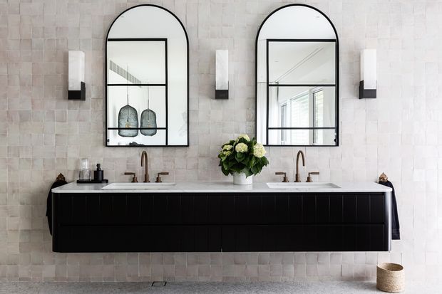 A refined double vanity bathroom with a long wall-mounted black vanity, two white basins, brass-toned tapware, arched black-framed mirrors, and matching wall sconces. The walls are clad in pale textured rectangular tiles, with a small floral arrangement on the vanity and a pale stone or concrete-look floor. The mirrors reflect a bright adjoining space and pendant lights.