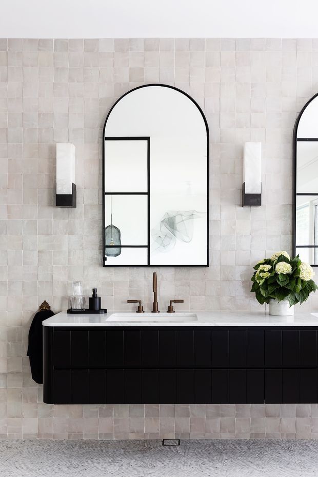 A refined contemporary bathroom vanity with a long black floating cabinet, white integrated countertop and basin, brushed brass tapware, arched black-framed mirror, and twin wall sconces. The walls are clad in pale handmade-look square tiles, with a textured light grey floor and restrained decorative styling.