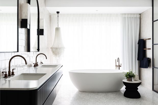 Bright, refined contemporary bathroom with a double vanity, arched brushed-metal tapware, white tiled walls, a freestanding oval bathtub, sheer full-height curtains, and dark timber or black cabinetry. A sculptural pendant and black side table with greenery add decorative contrast.