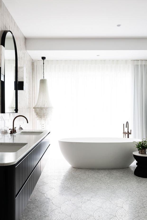 Bright, refined bathroom with a freestanding white bathtub positioned in front of sheer floor-to-ceiling curtains. A long dark vanity with twin integrated basins and brushed metal fixtures runs along the tiled wall, complemented by a large arched black-framed mirror and a decorative pendant light. The floor features a small-scale light mosaic tile pattern, while the room is accented with black and white finishes and abundant natural light.