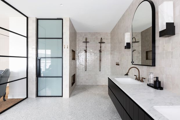 Bright contemporary bathroom with a large double vanity, white stone benchtop, dark cabinetry, arched black-framed mirror, brass tapware, pale tiled walls, and a walk-in shower. A black-framed glazed partition provides a view into an adjacent room.