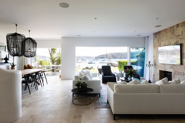 Bright open-plan coastal living and dining area with large windows overlooking a marina or waterfront. The room features pale stone-look flooring, white furnishings, a white sectional sofa, a dark armchair, a stone-clad fireplace wall with television, a timber dining table with black chairs, and distinctive woven pendant lights.