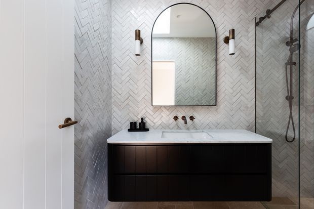 A refined contemporary bathroom featuring a floating dark timber vanity with an integrated pale benchtop, wall-mounted brass-toned tapware, an arched black-framed mirror, cylindrical wall sconces, and a frameless glass shower enclosure. The walls are clad in a light herringbone-patterned tile, creating a textured yet restrained finish.