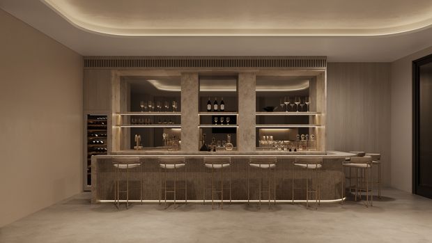 A sophisticated built-in home bar with extensive illuminated glass shelving, wine storage, bottles and glassware, a long stone-topped counter, and integrated bar stools. The room has warm taupe and brown finishes, ribbed cabinetry, a sculptural curved ceiling detail with concealed lighting, and a dark-framed doorway.