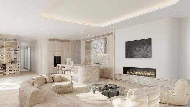 A bright, luxurious open-plan living room with a curved cream sectional sofa, sculptural cream lounge chair, glass coffee table, linear built-in fireplace, wall-mounted artwork, and a pale stone kitchen island in the background. The space features soft layered lighting, light wood and stone finishes, recessed ceiling detailing, and a calm neutral palette.