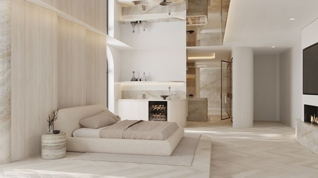 A warm, minimalist open-plan interior featuring a low upholstered bed or daybed in the foreground, pale timber wall panelling, integrated shelving and a fireplace. The space extends toward a compact kitchen or bar area and a glass-enclosed bathroom or shower zone, with a large wall-mounted television and another fireplace on the right. Soft beige, cream and light-wood finishes create a calm, luxurious atmosphere.