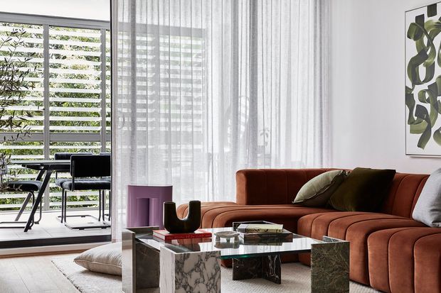 A refined contemporary living room with a rust-brown curved sectional sofa, marble-clad block coffee table with a glass top, and neutral flooring. Sheer curtains soften the full-height glazing, which opens to a balcony furnished with outdoor seating and screened by horizontal louvres. Minimal artwork and sculptural decor complete the space.