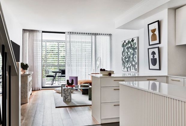 Bright open-plan interior viewed from the kitchen, with a white island featuring integrated cabinetry and a fluted front, pale timber flooring, and a living area beyond. Floor-to-ceiling sheer curtains frame glazed doors leading to a balcony or exterior. Contemporary abstract artwork, a neutral sofa, and modern furniture create a refined, airy scheme.