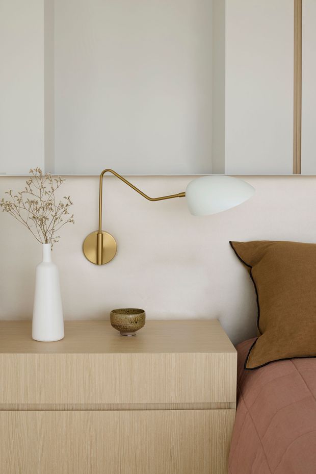 Close-up of a softly styled bedroom corner featuring a light timber bedside cabinet with integrated drawers, a white vase with dried stems, a small ceramic bowl, and a brass wall-mounted swing-arm lamp. A blush-pink upholstered bed with a warm brown cushion is visible at right. The palette is warm, muted, and minimalist.