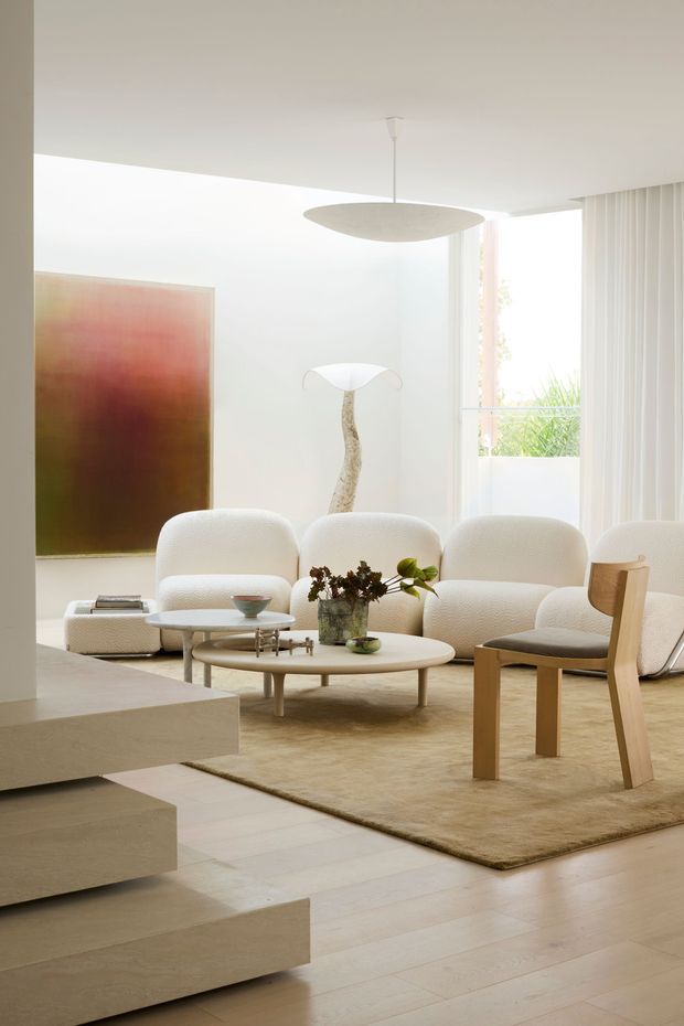 Bright, refined living room with a soft neutral palette, sculptural cream boucle seating, a low rounded coffee table, pale timber flooring, and a large beige area rug. Full-height glazing with vertical blinds brings in abundant daylight, while an abstract artwork, sculptural floor lamp, and suspended oval pendant add understated visual interest. Broad pale stone steps connect the living area to a lower level.