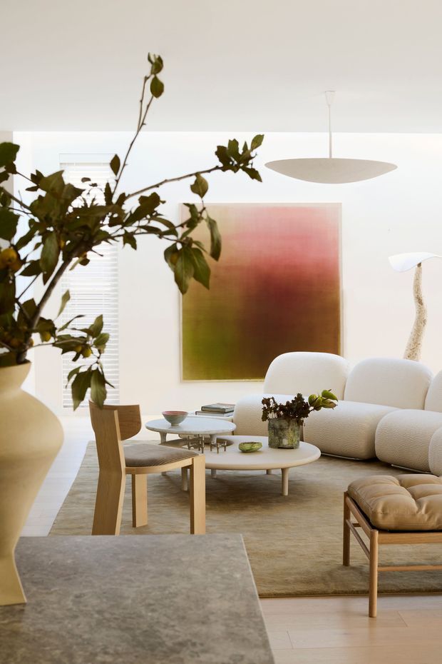 A bright, refined living room with a warm neutral palette, featuring a sculptural cream sectional sofa, light timber lounge chairs, round coffee tables, a textured area rug, and minimalist suspended lighting. A large abstract artwork and leafy branch arrangement add soft organic colour and visual interest. The space has white walls, pale timber flooring, and contemporary furnishings with rounded forms.