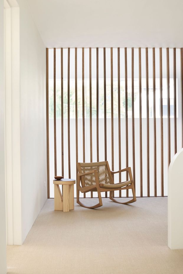 A bright, minimalist sitting area or hallway nook with a sculptural wooden rocking chair and small round side table on a pale carpet. The space features white walls and ceiling, a full-height window, and a striking vertical timber slat screen that filters light and provides visual separation. The restrained palette and natural wood create a calm, warm atmosphere.