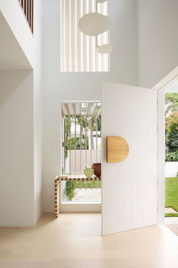 Bright double-height residential entryway with white vertical wall panelling, pale timber flooring, a large open pivot door, clerestory glazing, and sculptural pendant lights. A timber console with decorative objects sits in front of a garden-facing window, while a covered outdoor area and greenery are visible beyond.