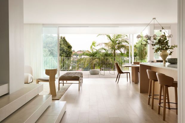 Bright, open-plan contemporary interior with a minimalist living and dining area connected to a large glazed balcony or terrace. The room features pale flooring and walls, a sculptural timber side table, upholstered lounge furniture, a dining table, and a kitchen or bar counter with timber stools. Full-height glazing and open balcony railings overlook lush greenery and palm trees, creating an airy indoor–outdoor connection.