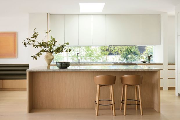 Bright contemporary kitchen centered on a large island with a light timber-clad waterfall-style front, pale stone benchtop, integrated light cabinetry, and two timber stools. A window splashback brings in garden views, while a skylight adds overhead illumination. A built-in dark banquette and framed artwork are visible at the left.