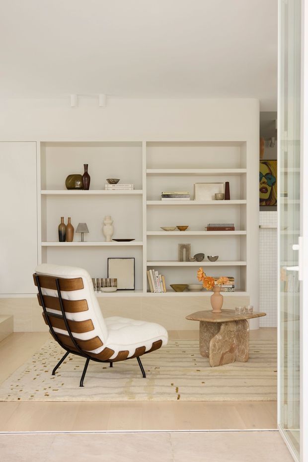 A refined, warm-toned living room with a built-in cream display wall featuring open shelving and curated decorative objects. A sculptural white-and-brown lounge chair sits on a pale patterned rug beside a rounded natural stone coffee table with a vase of flowers. Full-height glazed doors frame the right side, and a partial artwork and tiled area are visible beyond.