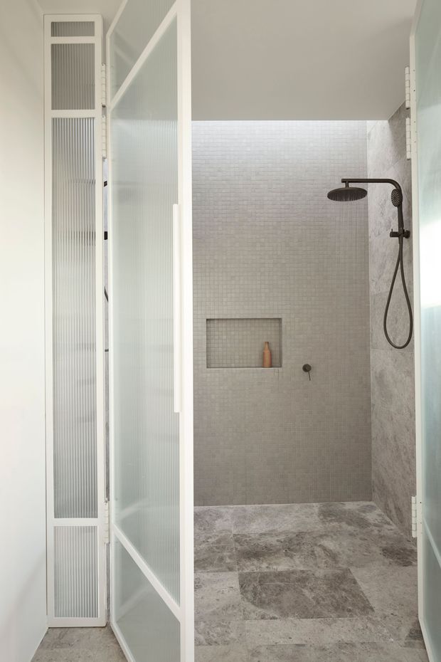 A bright, compact bathroom shower area viewed through open white-framed ribbed-glass doors. The walk-in shower features small pale mosaic wall tiles, larger mottled stone-look floor tiles, a recessed wall niche, and dark bronze or black shower fittings including a rain shower and handheld outlet. The overall design is clean, contemporary, and neutral with soft natural light.