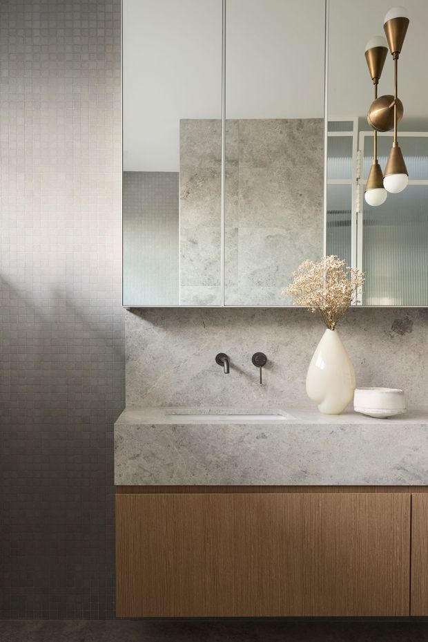 A refined contemporary bathroom vanity with a floating ribbed timber cabinet, pale stone benchtop and matching stone backsplash, dark small-format tiled wall, and mirrored wall cabinets. Brushed brass sculptural pendant lights and a white ceramic vase add warm decorative accents. The composition is clean, softly lit, and luxurious.
