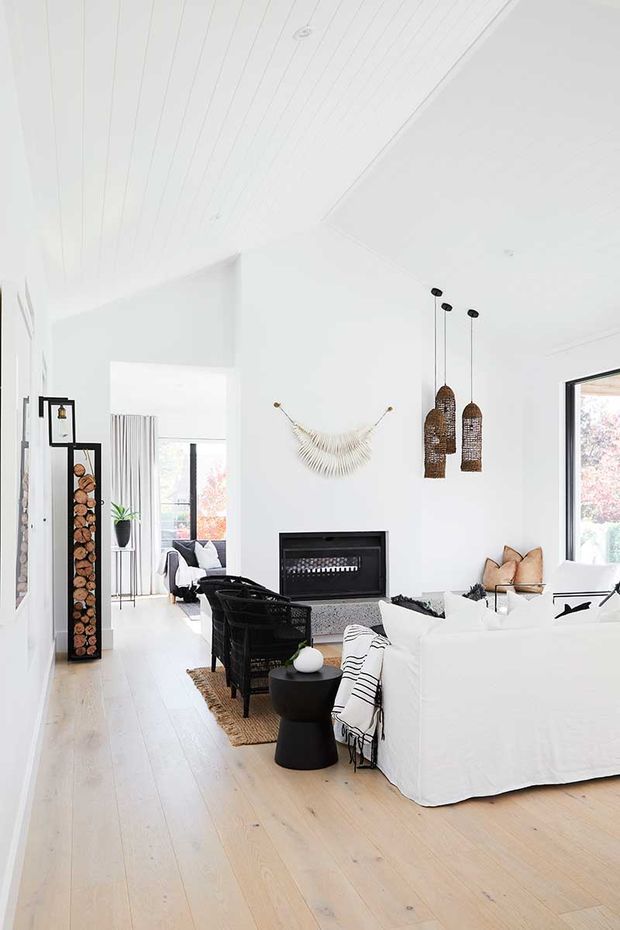 Bright, airy open-plan living room with white walls and ceiling, pale timber flooring, a large white sectional sofa, black lounge chairs, and a dark built-in fireplace. Woven pendant lights, a sculptural wall hanging, timber log storage, and expansive windows add warm natural texture and contrast.