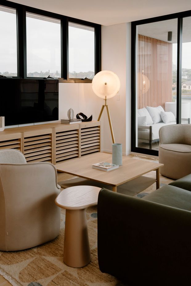 A refined contemporary living room with large black-framed windows and glazed doors, a low timber entertainment unit with slatted cabinetry, a rectangular timber coffee table, sculptural upholstered lounge chairs and sofa, and a warm tripod floor lamp. Neutral beige, timber and black tones create a calm, softly layered interior. A bedroom or lounge area is visible through the glazed door, with vertical timber wall battens and water or landscape beyond.