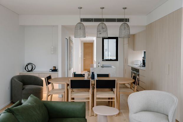 Bright open-plan apartment interior combining a dining area, compact kitchen and living room. A timber dining table with chairs sits beneath three woven pendant lights, with pale timber full-height cabinetry along the right wall. A green sofa and upholstered lounge chairs occupy the foreground, while a small window and concealed air-conditioning grille appear at the rear. The overall look is calm, contemporary and warm with light neutral finishes and natural timber.