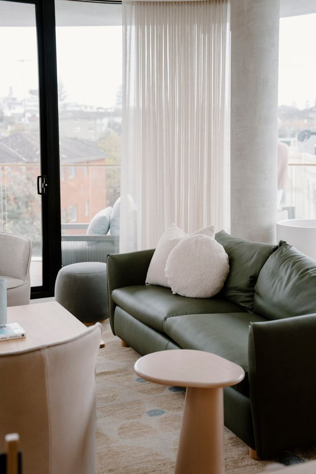 A bright, contemporary living room with a deep olive-green sofa, cream boucle cushions, pale upholstered chairs, light timber side tables, and a softly patterned neutral rug. Full-height sheer curtains and black-framed glazing open to an urban outlook, while a rounded pale column adds architectural interest.