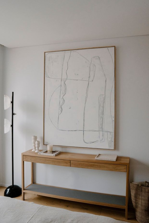 A minimalist interior vignette featuring a slim natural timber console table with a grey lower shelf, set against a white wall. A large abstract line artwork in a simple timber frame hangs above the console, with small neutral decorative objects and books on top. A black floor lamp, woven basket, timber flooring, and the edge of a light upholstered seating piece are also visible.