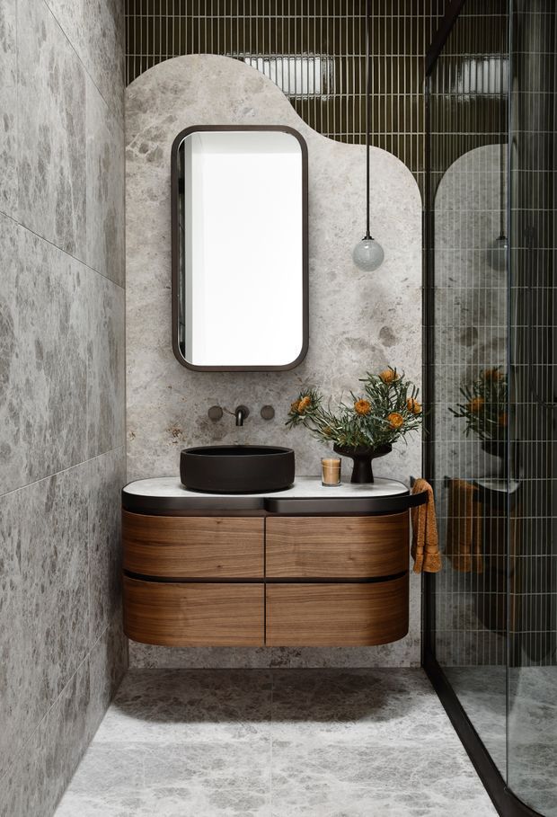 A sophisticated contemporary bathroom vanity area featuring a rounded-edge wall-mounted timber vanity with a dark vessel basin and pale stone benchtop. Large-format grey veined stone tiles cover the wall and floor, while a sculptural stone feature panel frames the rounded mirror. A glass shower screen with dark framing, a suspended globe pendant, and restrained botanical styling complete the space.