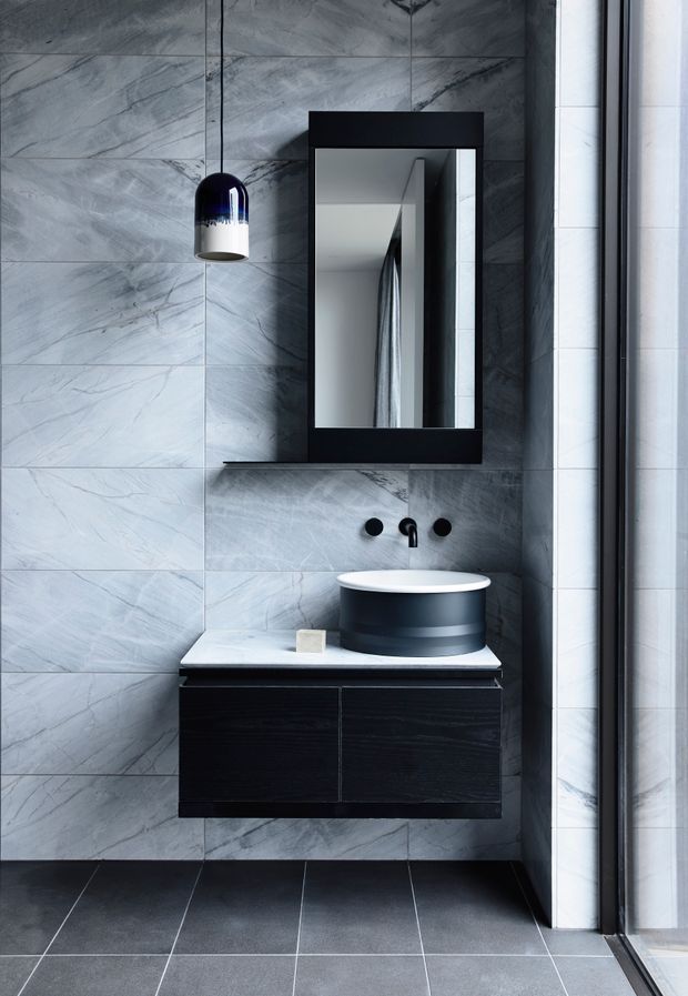 A compact contemporary bathroom with large grey marble-look wall tiles, dark charcoal floor tiles, a floating black timber vanity with a pale stone benchtop, round vessel basin, matte black tapware, and a black-framed rectangular mirror. A dark pendant light hangs beside the mirror, while a glazed shower or window enclosure is visible at the right.