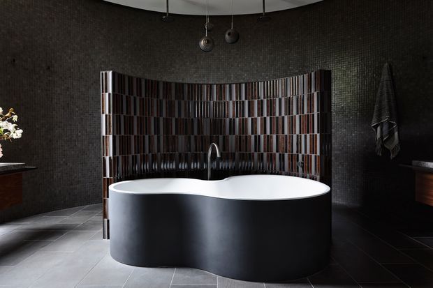 A dramatic, dark-toned bathroom featuring a sculptural freestanding double-ended bathtub with a matte black exterior, set against black mosaic walls and a curved vertical feature-tile backsplash. A central wall-mounted or floor-mounted bath tap rises behind the tub, with dark tiled flooring, a curved ceiling detail, pendant lighting, and dark timber cabinetry at the sides.