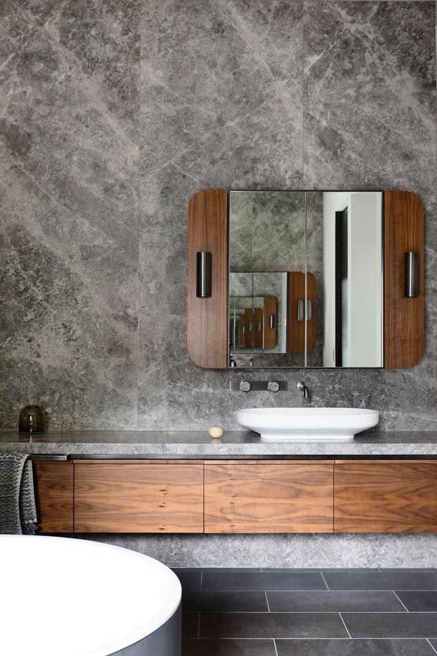 A sophisticated contemporary bathroom featuring a floating walnut vanity with integrated drawers, a dark stone benchtop, white vessel basin, wall-mounted tapware, and a large rounded-corner mirrored cabinet. The walls and floor are finished in richly veined grey stone or porcelain, with a freestanding white bathtub partially visible in the foreground. The overall scheme is moody, refined, and material-focused.