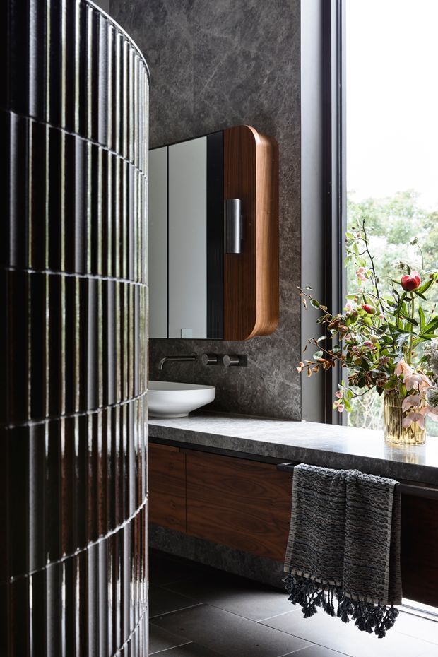 A refined contemporary bathroom featuring a floating dark timber vanity with a pale stone benchtop, white vessel basin, wall-mounted dark tapware, and a rounded timber-framed mirrored cabinet. The room has dark stone wall cladding and large dark floor tiles. A curved ribbed black-and-metal partition partially screens the vanity, while a tall window brings in views of greenery and natural light.