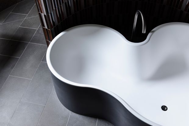 Close-up view of a sculptural freestanding bathtub with a flowing asymmetrical double-lobed form, finished in white inside and dark charcoal outside. It sits on large-format grey floor tiles beside a dark glossy vertically textured tiled wall, with a minimalist curved chrome tap visible behind it.
