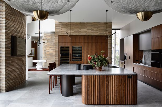 Open-plan contemporary kitchen and dining area with a large pale stone island featuring a vertically slatted timber-clad base, dark wood cabinetry, integrated appliances, and a built-in wine storage wall. A curved exposed brick feature wall with a television defines the adjacent living area. The space has sculptural mesh pendant lights, pale stone flooring, warm timber finishes, and abundant natural light.