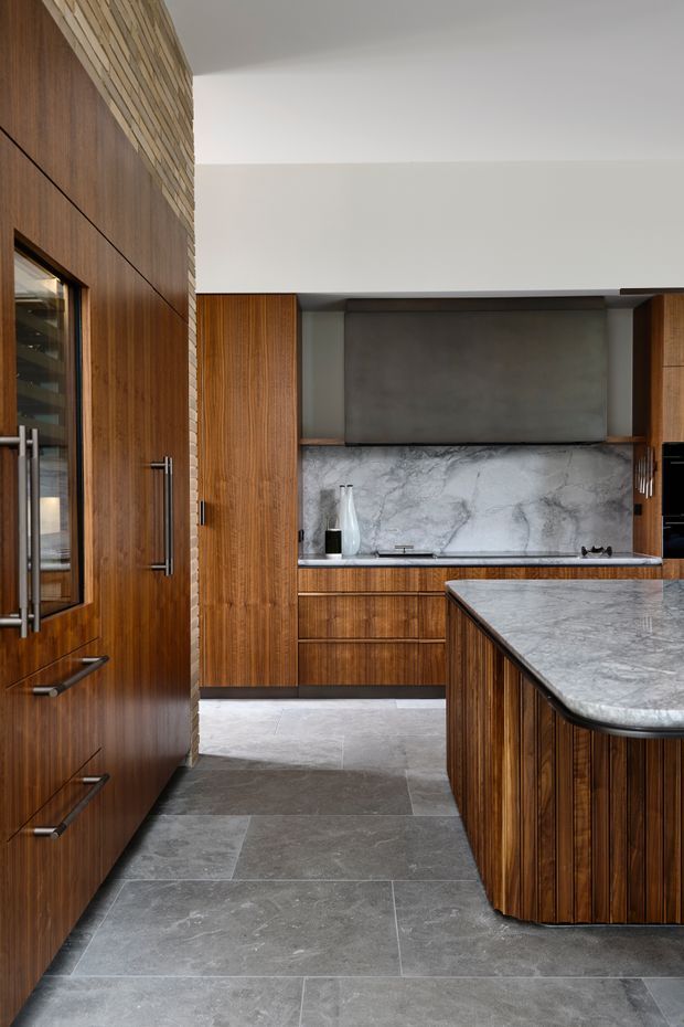 A refined contemporary kitchen with extensive dark timber cabinetry, a large stone island with rounded edges, grey stone flooring, a dramatic marble-look splashback, and a dark metal rangehood surround. The composition also shows a tall timber-clad wall and integrated appliances, creating a warm, luxurious, material-focused interior.