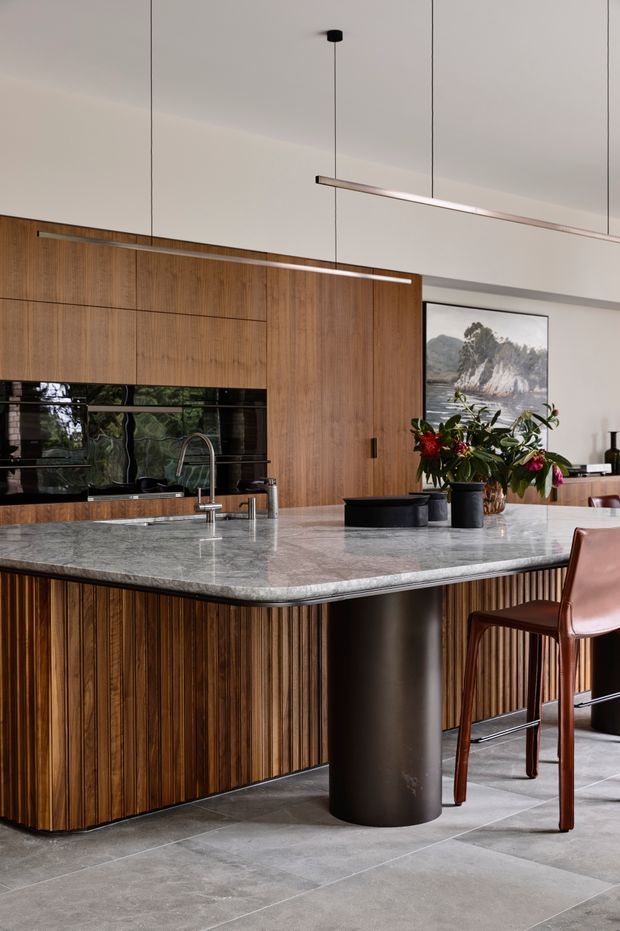 A refined contemporary kitchen featuring extensive warm timber cabinetry with vertical fluted detailing, a large grey marble island benchtop with a rounded edge, dark cylindrical island supports, integrated black appliances, and a sculptural linear pendant light. A dining chair, floral arrangement, artwork, and dark grey tiled flooring complete the sophisticated interior.