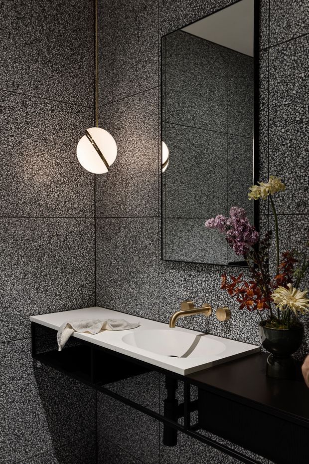 A dramatic contemporary bathroom vanity with a sculptural white integrated basin set into a black floating console. The walls are clad in dark, small-format textured pebble-like panels with visible grid joints, complemented by a large frameless mirror, brushed brass wall-mounted tapware, and a spherical pendant light. Dark floral arrangements provide contrast against the moody monochromatic finish.