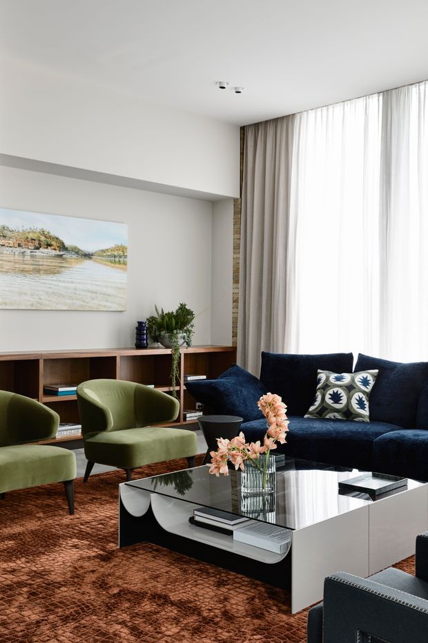 A refined living room with a deep navy velvet sofa, olive-green lounge chairs, a glossy black-and-white rectangular coffee table, and a textured rust-brown rug. A timber built-in shelving and media console runs along the rear wall beneath a landscape artwork, while full-height sheer and taupe curtains soften the bright window wall. A narrow exposed brick or stone vertical feature adds texture beside the curtains, and floral arrangements provide a small decorative accent.