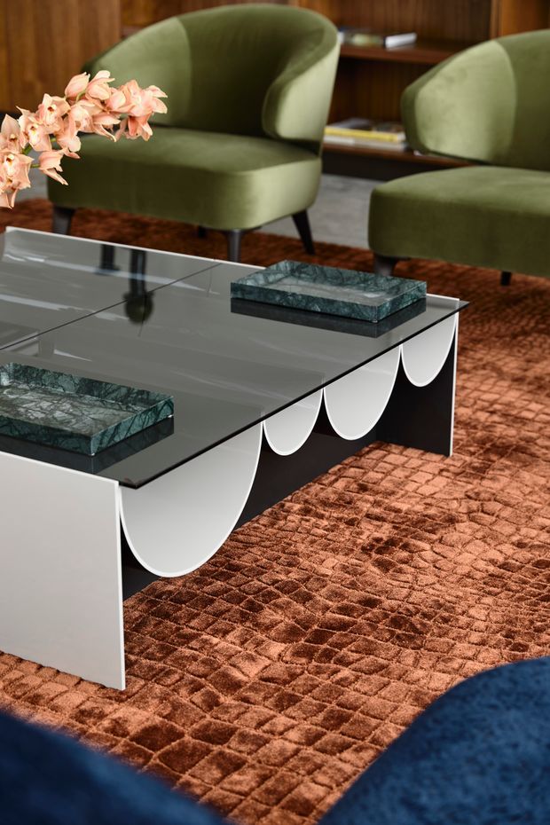 A richly styled living room vignette centered on a sculptural rectangular coffee table with a glossy dark stone or glass top, white side panels, and repeated semicircular decorative forms along its base. Olive-green upholstered lounge chairs sit behind it, with a dark timber wall and shelving in the background. A rust-brown textured rug, pink flowers, and green stone accessories add warm, luxurious colour and tactile contrast.