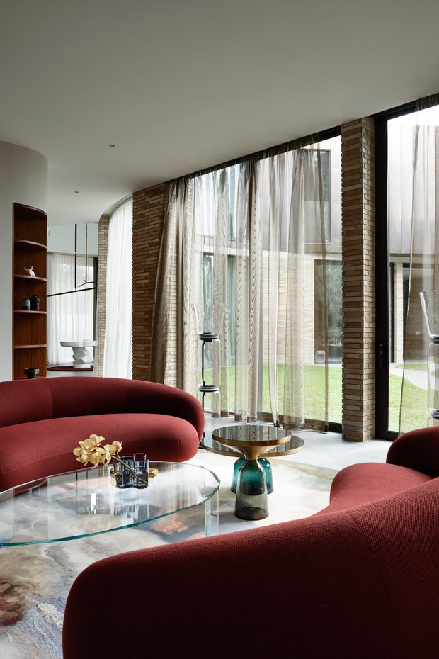 A refined living room with sweeping floor-to-ceiling glazing, sheer curtains, warm exposed brick columns, and curved burgundy upholstered sofas arranged around a large glass coffee table. A sculptural side table, built-in timber shelving, and a glimpse of an adjoining dining or workspace area complete the sophisticated interior, with a landscaped courtyard visible beyond the windows.