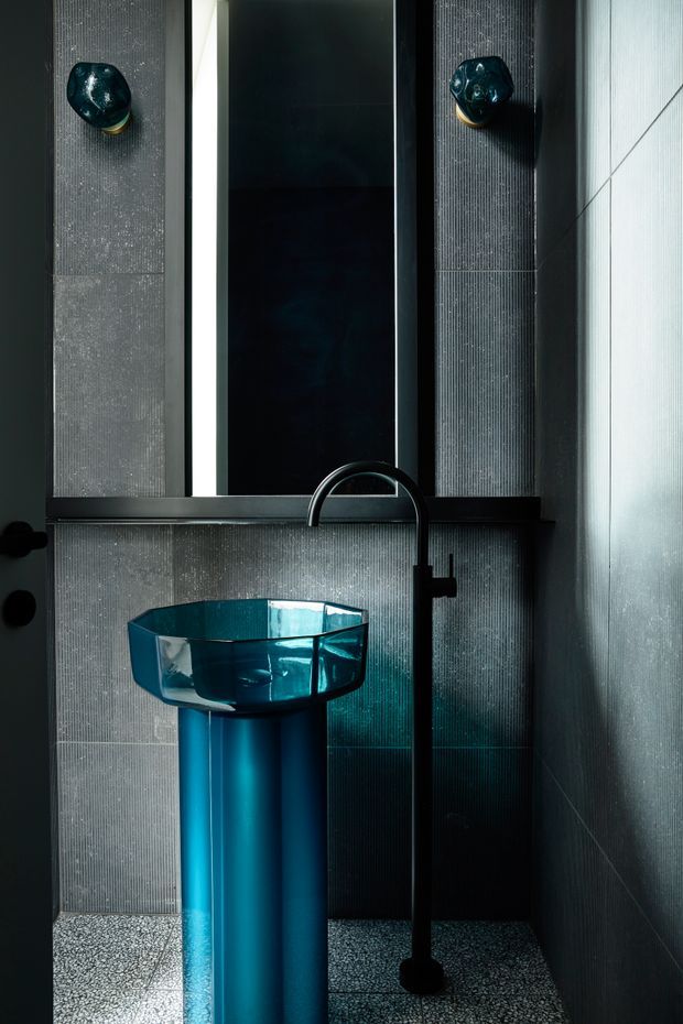 A dramatic, compact bathroom featuring a sculptural translucent turquoise pedestal basin with a tall matte black gooseneck tap. Dark vertically textured wall tiles surround a narrow mirror, while terrazzo-style flooring and turquoise-black accents create a moody, jewel-toned interior.