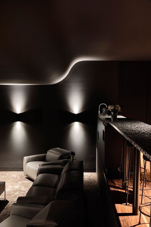 A moody, high-end residential interior combining a dark living area with a bar or kitchen counter. Sculptural integrated wall lighting creates dramatic pools of light across dark walls, while a large pale sectional sofa and lounge chair sit on a light textured carpet. The counter has a dark patterned stone surface, dark cabinetry, and slim metal bar stools over a warm timber floor. The ceiling features a curved, illuminated transition that adds a distinctive architectural element.