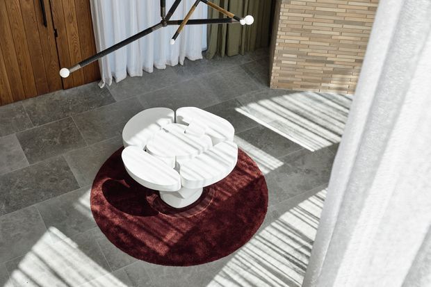 Sunlit contemporary interior with large grey stone floor tiles, a round deep burgundy rug, sculptural white circular coffee table, timber door or wall panelling, sheer curtains, and a distinctive linear pendant light. A pale horizontal brick feature wall and strong striped sunlight add texture and contrast.